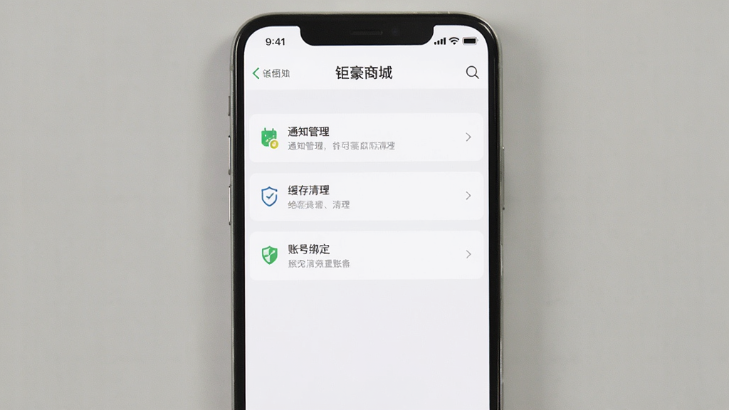 一张展示钜豪商城APP设置页面的图片,画面中央是手机屏幕,屏幕上显示通知管理、缓存清理、账号绑定等设置选项,布局清晰,风格简洁,色彩以白色和浅灰色为主,画质干净,无多余元素。