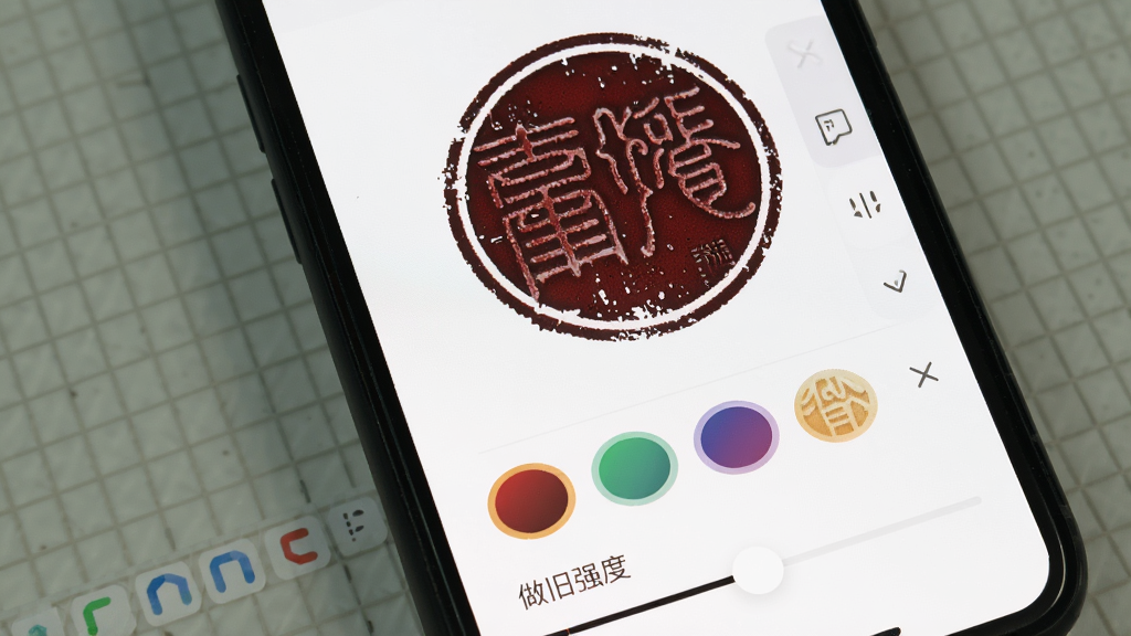 手机屏幕显示印章设计师App的颜色调节界面,一枚圆形印章呈现暗红色并带有轻微做旧斑驳效果,屏幕下方有颜色选择圆圈和「做旧强度」滑块,背景是白色工作区,界面布局清晰,风格简洁,画质干净,无多余元素