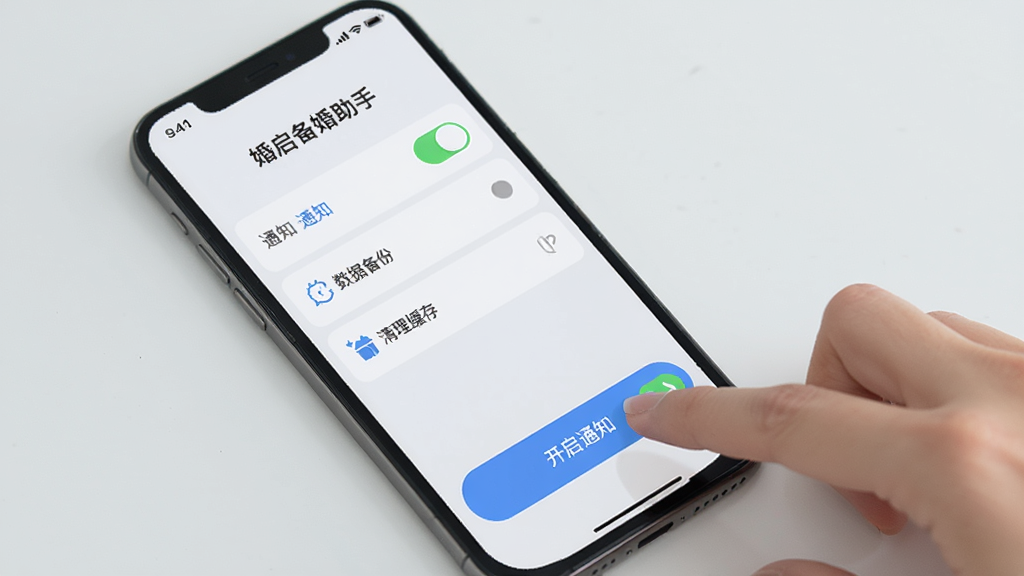 一张展示手机屏幕上婚启备婚助手App设置界面的图片,界面中有通知开关、数据备份、清理缓存等选项,旁边有一只手正在点击“开启通知”按钮。风格简洁,图标清晰,背景是浅灰色,画质干净,无多余元素。