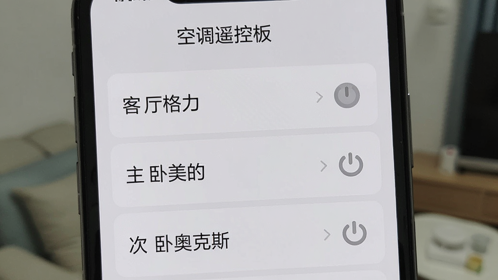 一张展示手机屏幕的图片,屏幕上打开着“空调遥控板”的设备列表页面,列表中显示三个已保存的空调设备,分别标注为“客厅格力”“主卧美的”“次卧奥克斯”,每个设备旁边有一个电源开关小图标,界面背景为浅灰色,文字清晰,布局整齐,风格简约现代,画质干净无多余元素