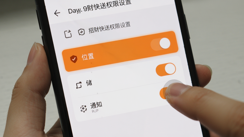 手机屏幕展示招财快送权限设置界面,包含位置、存储、通知三个开关选项,旁边有手指点击开启位置的示意,界面风格简洁,橙色高亮,背景为手机设置页面,画质干净无多余元素