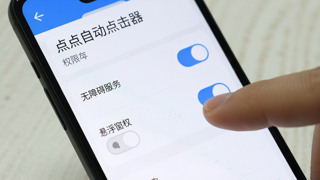 手机屏幕上显示“点点自动点击器”的权限设置页面,页面中有无障碍服务开关和悬浮窗权限开关,用户的手指正在点击开关按钮,界面风格简洁,以白色和蓝色为主,画质清晰,无多余元素