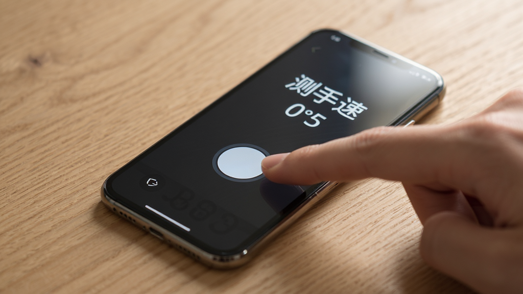 手机放在桌面上,一根手指正在点击屏幕上的圆形按钮,屏幕显示“测手速”和毫秒数,背景是木质桌面,光线柔和,风格写实,细节清晰,画质干净,无多余元素