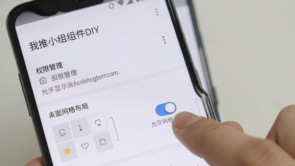 一部安卓手机屏幕显示“我推小组件DIY”的设置页面,页面中有权限管理选项和桌面网格布局选择项,画面中有一只手正点击“允许显示在其他应用上层”的开关按钮,界面风格简洁,文字清晰,画质干净,无多余元素