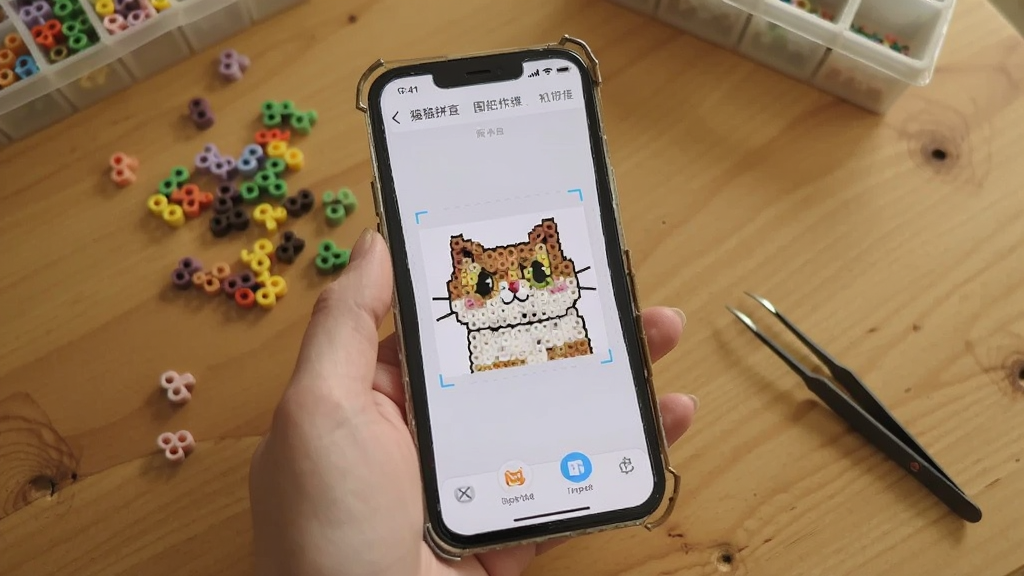 一张展示豆猫拼豆新手操作场景的图片,画面中一只手拿着手机,屏幕上显示着豆猫拼豆App的图片导入转图纸界面,旁边桌面上散落着几颗彩色拼豆和一把镊子,画面风格温馨日常,光线自然,细节清晰,画质干净,无多余元素