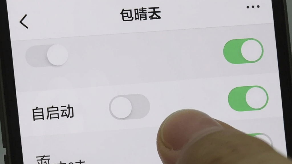一张手机截图,显示“包晴天”应用的设置页面,页面上有一些开关选项,背景为浅灰色,其中一项“自启动”开关被手指按住关闭,手指特写,画面干净,重点突出,画质清晰,无多余元素。