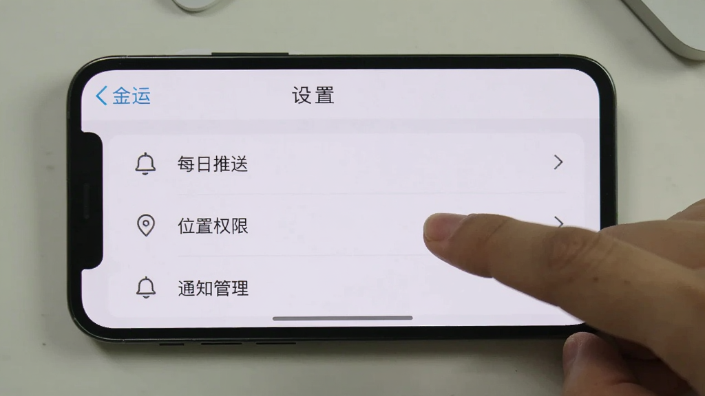 一张手机横屏截图,屏幕上显示“金运”APP的“设置”页面,页面中有“每日推送”、“位置权限”、“通知管理”等选项,旁边有一只手正在点击屏幕,背景是简洁的桌面,风格写实,画质干净,无多余元素