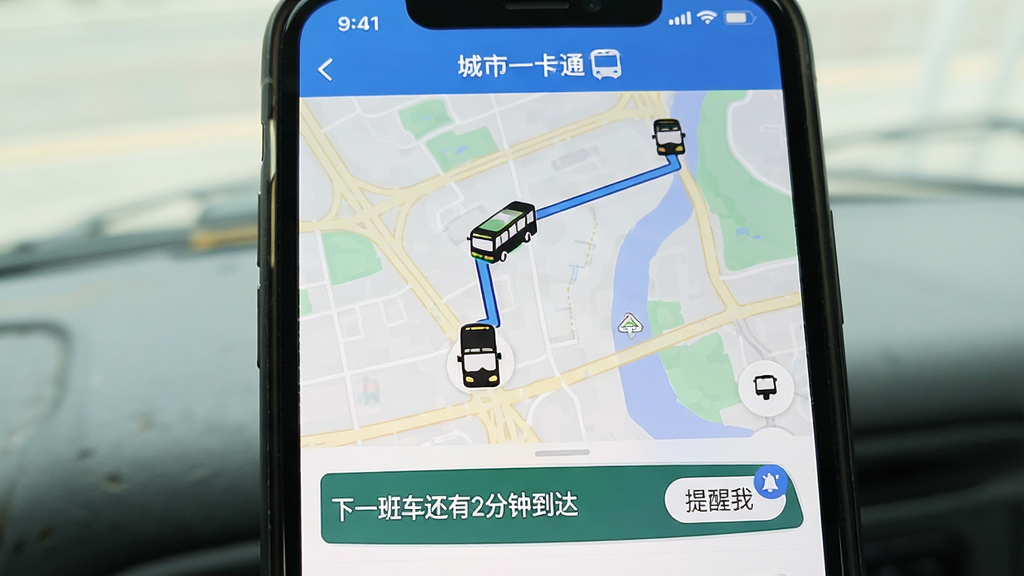 手机屏幕显示“城市一卡通”的实时公交页面,地图上清晰标注了公交车位置和行驶路线,底部列表显示“下一班车还有2分钟到达”,旁边有一个“提醒我”的按钮,画面干净整洁,信息直观易懂,无多余元素