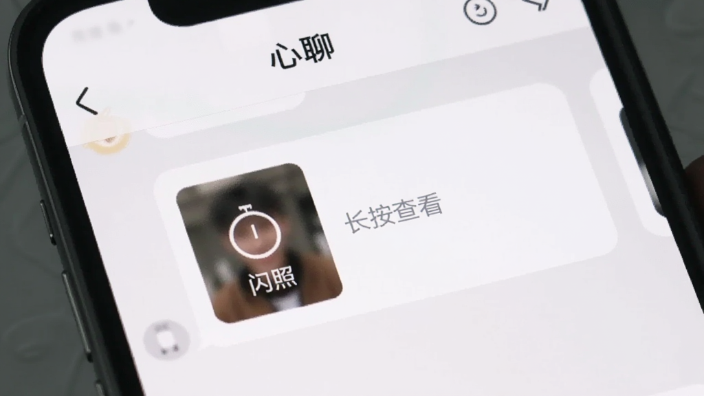 一个手机屏幕的局部特写,展示“心聊”的聊天界面,一条消息是一个模糊的图片缩略图,上面有一个倒计时图标和“闪照”字样,旁边有提示“长按查看”,界面设计简洁现代