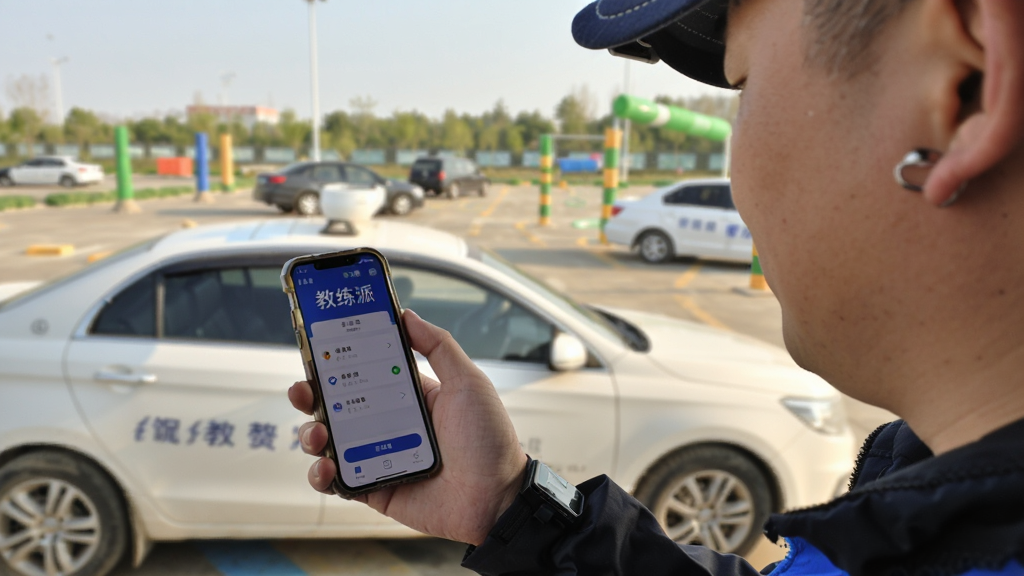 一位驾校教练在训练场边用手机打开教练派App查看学员签到记录,背景是教练车和训练场地,阳光充足,画面风格真实自然,手机屏幕上