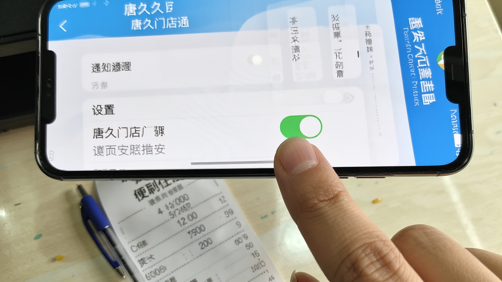 手机屏幕特写,显示“唐久门店通”的设置页面和通知管理界面,画面中有手指正在点击通知开关,背景是桌面上的便利店收银小票和圆珠笔,布局以上下结构呈现,上方是App界面,下方是桌面物品,风格为写实生活记录,色彩明亮自然,细节上手指动作清晰、界面文字可辨,画质干净无多余元素