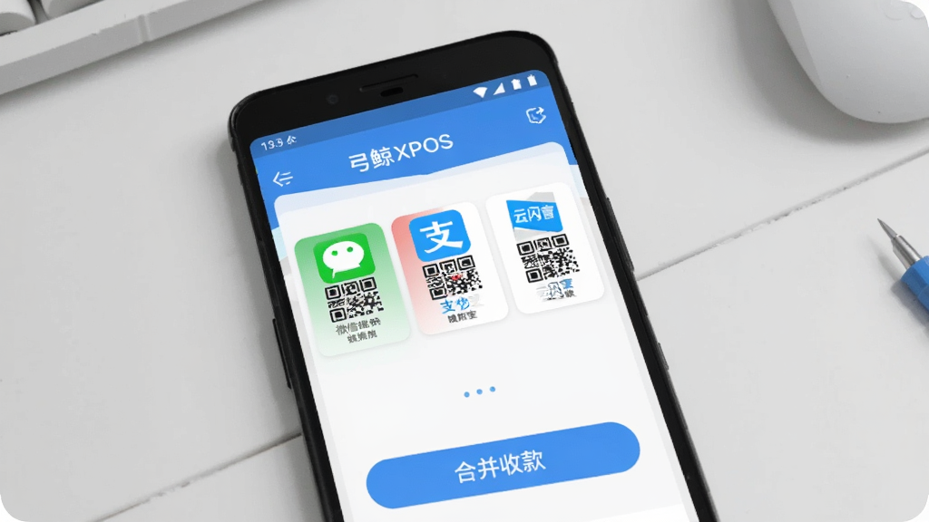 弓鲸XPOS 多场景收款码聚合 功能展示图,画面主体是一部安卓手机,屏幕内弓鲸XPOS应用界面显示微信、支付宝、云闪付三个收款码图标并排排列,下方有合并收款按钮,背景是简洁的浅灰色工作台,风格扁平化,画质清晰,无杂乱元素