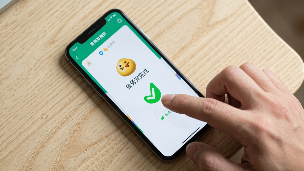 主体是一部手机放在桌面上,屏幕显示金豆荚的任务完成页面,旁边有一只手正在点击屏幕。布局是俯视视角。风格是生活化写实照片。细节上手指有自然的皮肤纹理,屏幕显示绿色对勾。画质清晰,背景是浅色木纹桌面,无多余元素。