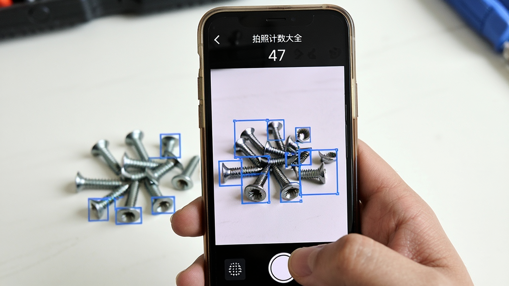 拍照计数大全软件操作界面特写,画面中一只手拿着手机对着桌上的螺丝钉拍照,手机屏幕里显示螺丝钉被蓝色方框逐个标注,屏幕上方显示“拍照计数大全”和计数结果“47”,主体是手机和螺丝钉,布局为第一视角拍摄,排版清晰,风格真实生活化,细节显示方框精准贴合螺丝钉边缘,画质明亮,画面干净,无多余元素