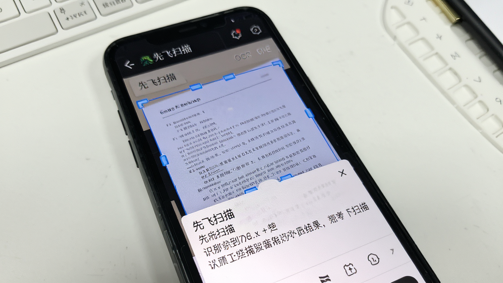 先飞扫描App的OCR文字识别界面,手机屏幕显示“先飞扫描”文字,屏幕中一张纸质文档图片上覆盖着蓝色识别框,下方弹出识别结果文本框,背景为白色工作台面,风格干净简约,画质清晰无多余元素